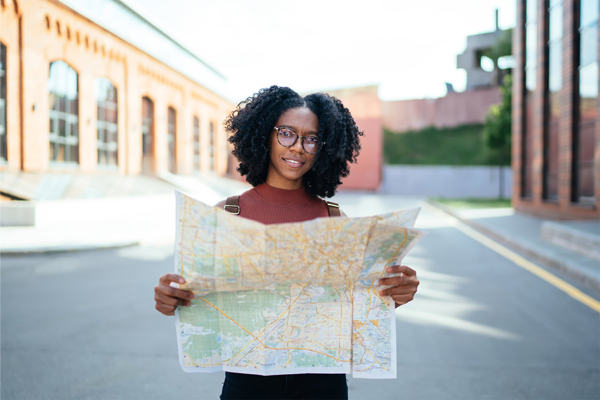 Black 30-year-old woman outside of two buildings wearing a backpack and glasses looking at a map. 