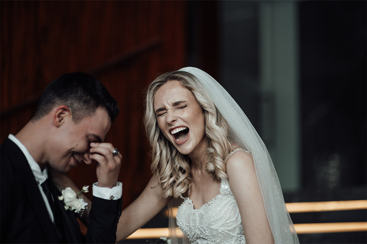 A young white couple on their wedding day are laughing with each other, while the man is pinching is nose with his fingers.