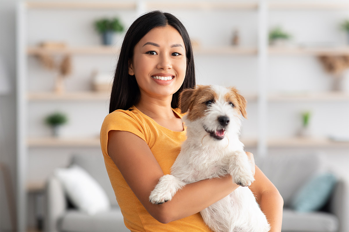 Asian 30-year-old woman in her living room smiling, wearing a yellow shirt and holding her dog. 