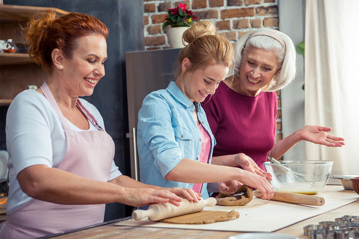 Three white women of different generations smile as they roll out dough on a kitchen table surrounded by baking tools and supplies 