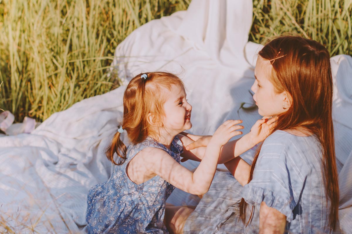 White red-headed toddler girl smiles and reaches in to tickle 5 year old white red headed sister as they sit on a white blanket in the grass on a sunny day