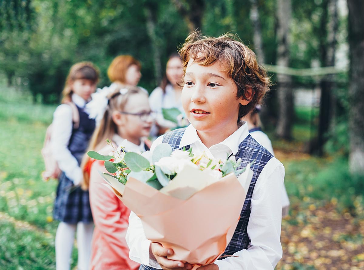 White little boy in a suite holding a bouquet of flowers outside, with groups of kids in the background