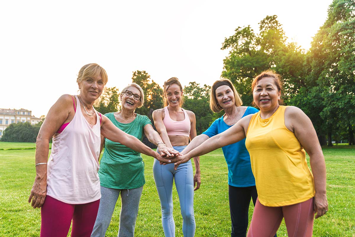 Five white women in their 50s to 60s wearing athletic attire smile outdoors during a sunny day with their hands in a pile 