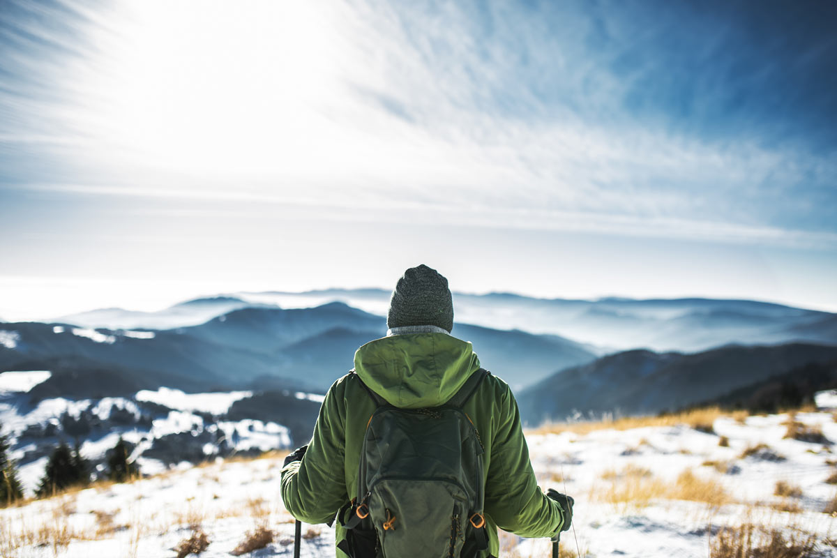 Man is hiking overlooking mountains wearing a green jacket, backpack, and beanie.