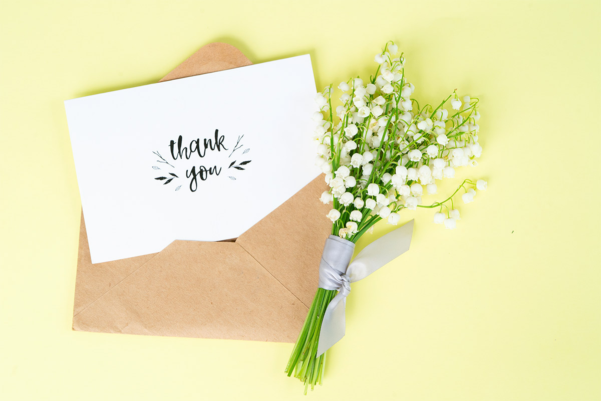 White thank you card pokes out of a brown envelope laying next to a small wrapped bouquet of small white flowers against a pale yellow backdrop 