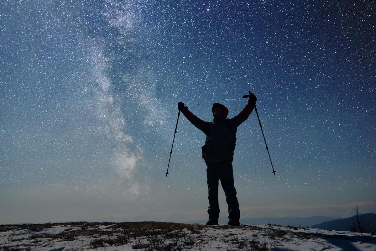 A silhouette of a skier lifts their arms up in front of a starry blue sky.