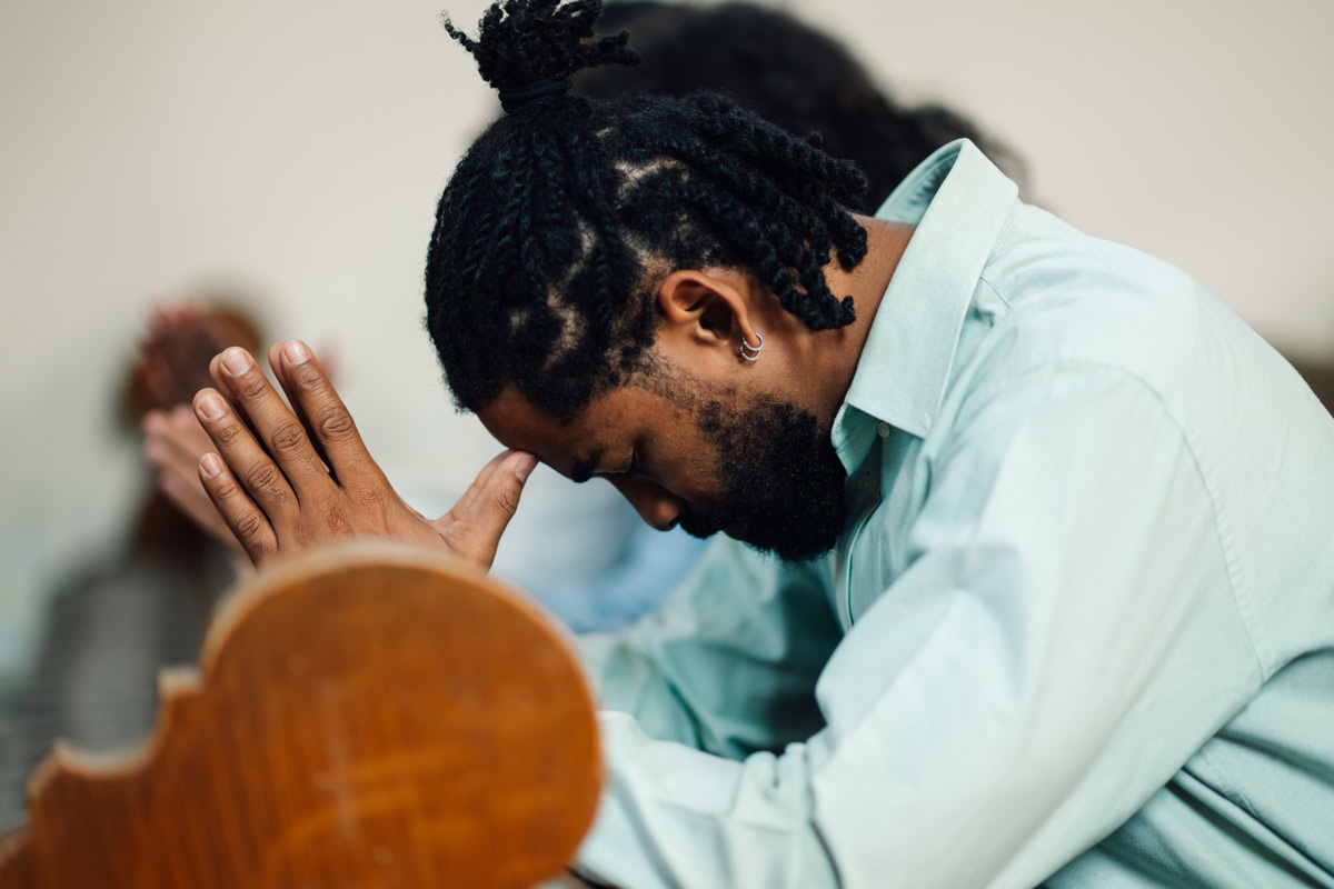 A black man sitting in a church pew bows his head and presses his hands together in prayer.