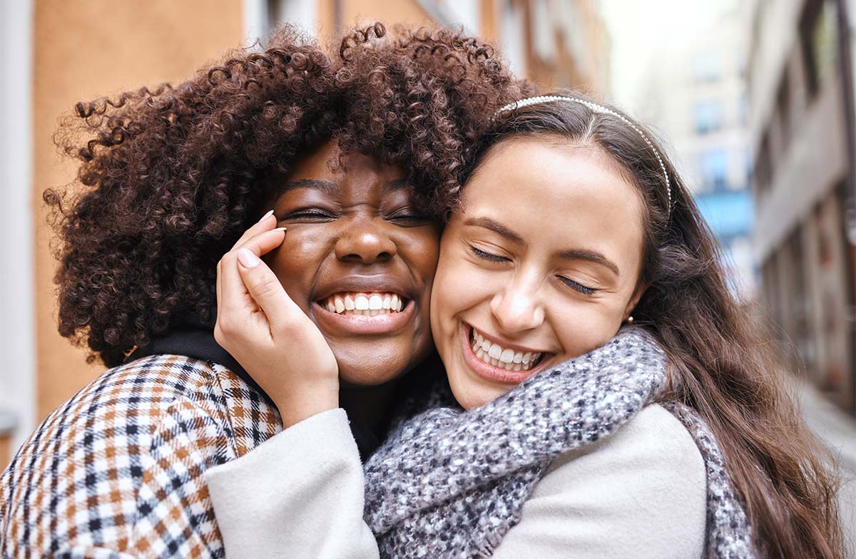 Two multiracial female friends in their 20s-30s hug close and share smiles with closed eyes