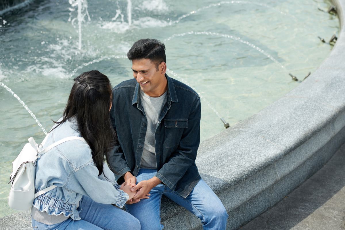 30-something Indian male holding woman’s hands while sitting at a fountain smiling 