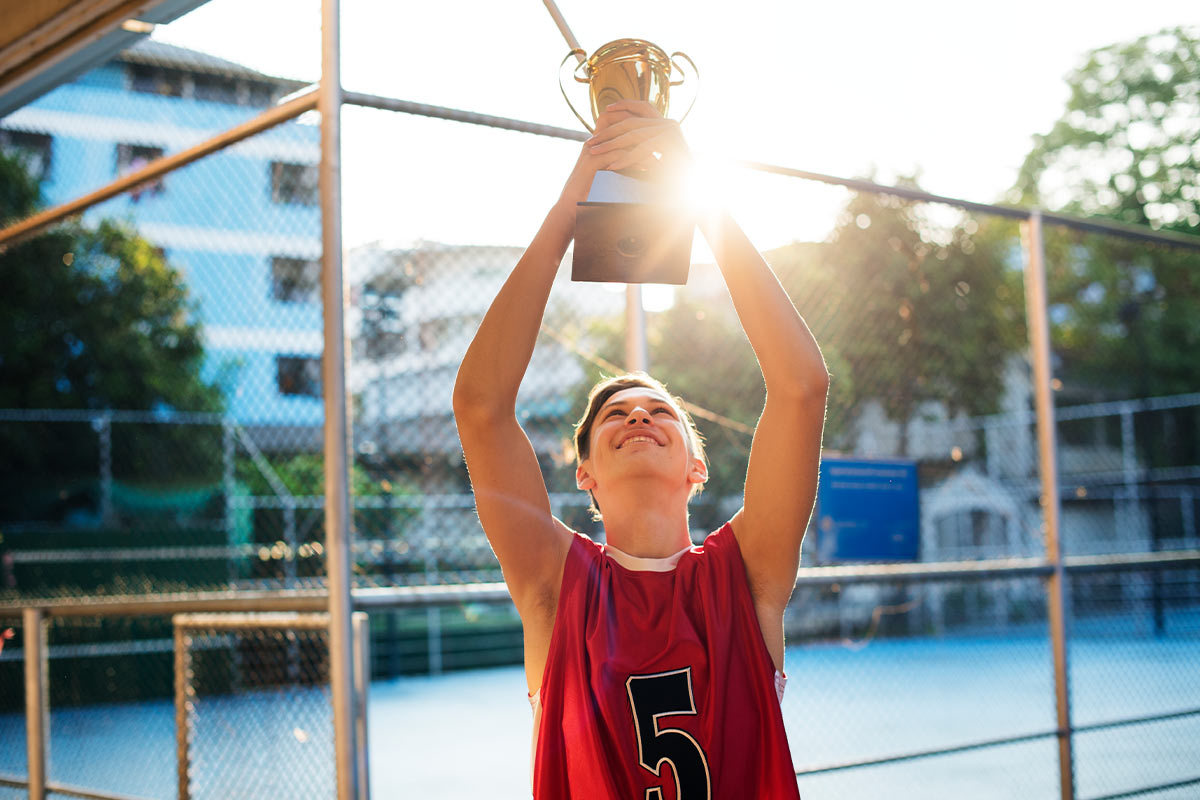 teenage white boy in a jersey holding a trophy outside on an outdoor court