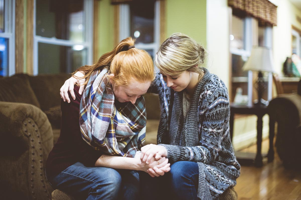 Two white women in their 20s bow their heads and hold hands in prayer while sitting in a living room