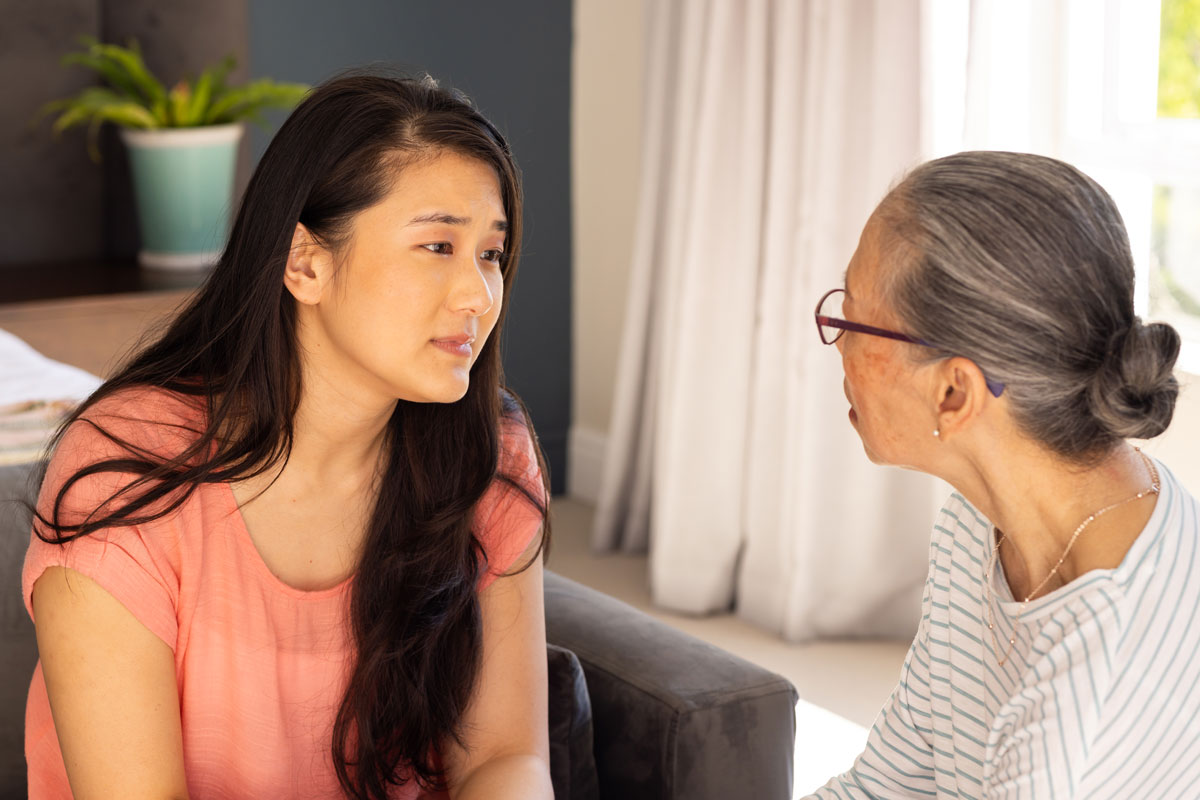 Asian 35-year-old daughter speaking to Asian 70-year-old mother on a couch in front of a window.