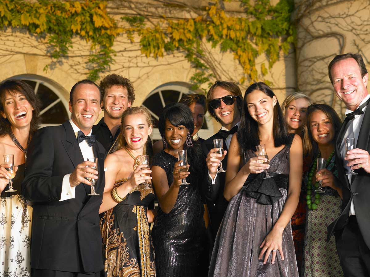 group of 11 people of multiple races and ages wear fancy attire and hold glasses while they smile for a group photo in front of a tan building with ivy growing on it  