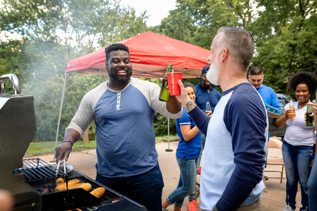 Black and white man, who are both middle aged, clinking drinks together by the grill outside, while black man is flipping burgers