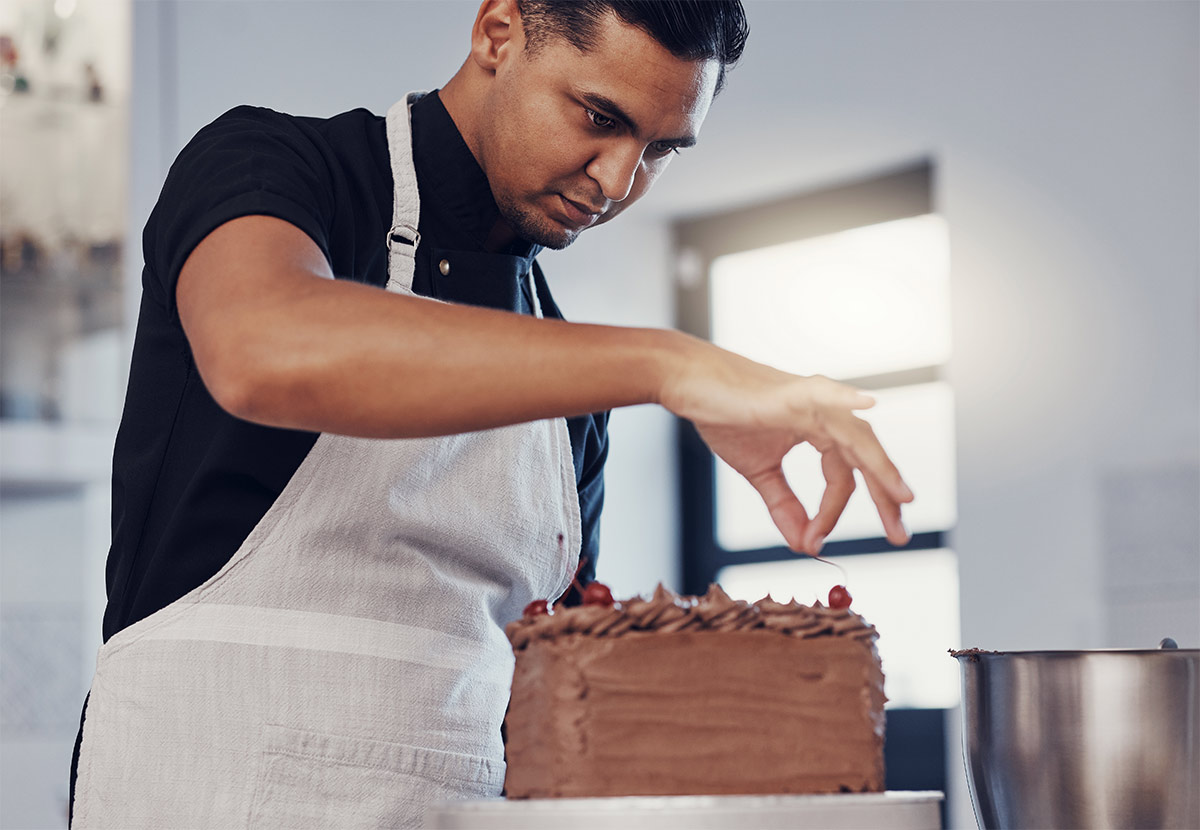 Hispanic man wearing an apron placing finishing touches on a chocolate cake with cherries on top. 