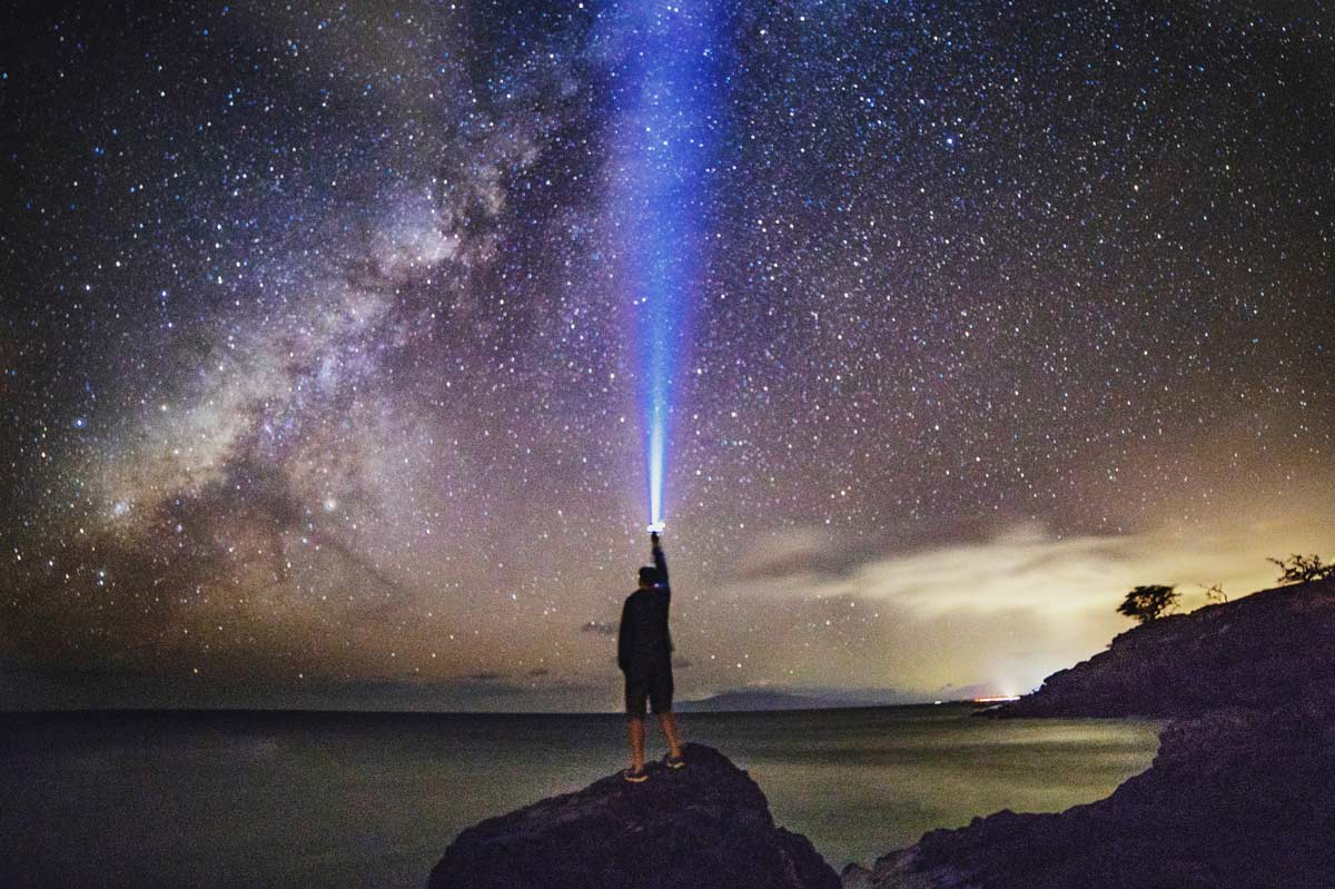 A person stands on a rock at night, pointing a bright beam of light towards the sky, illuminating the stars.