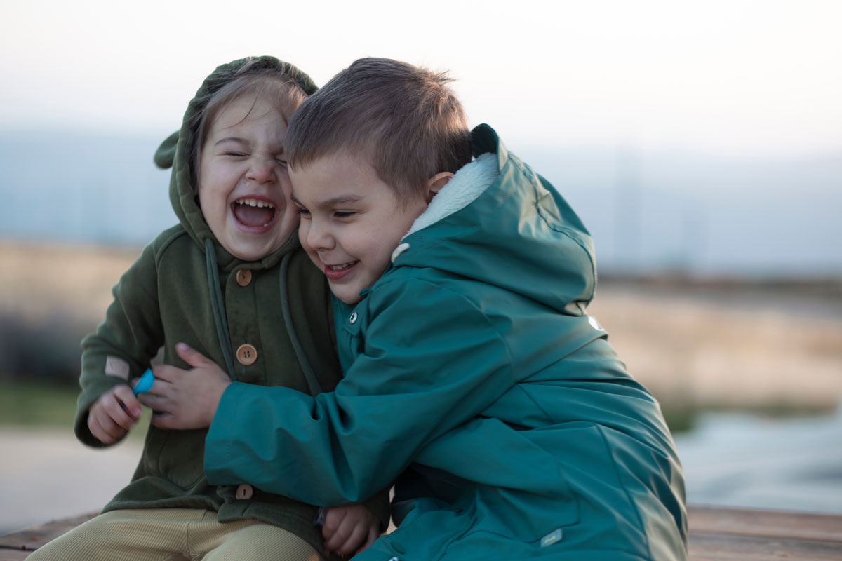 2 and 5 year old white brothers sit outside holding each other and laughing.