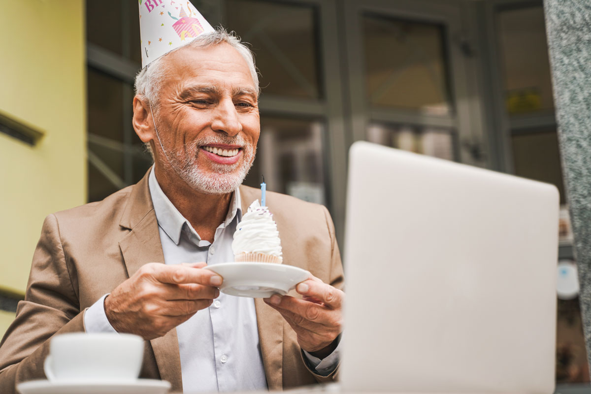 Older light skinned man smiling with a party hat holding a cupcake on a plate, he is smiling at his laptop at a café.