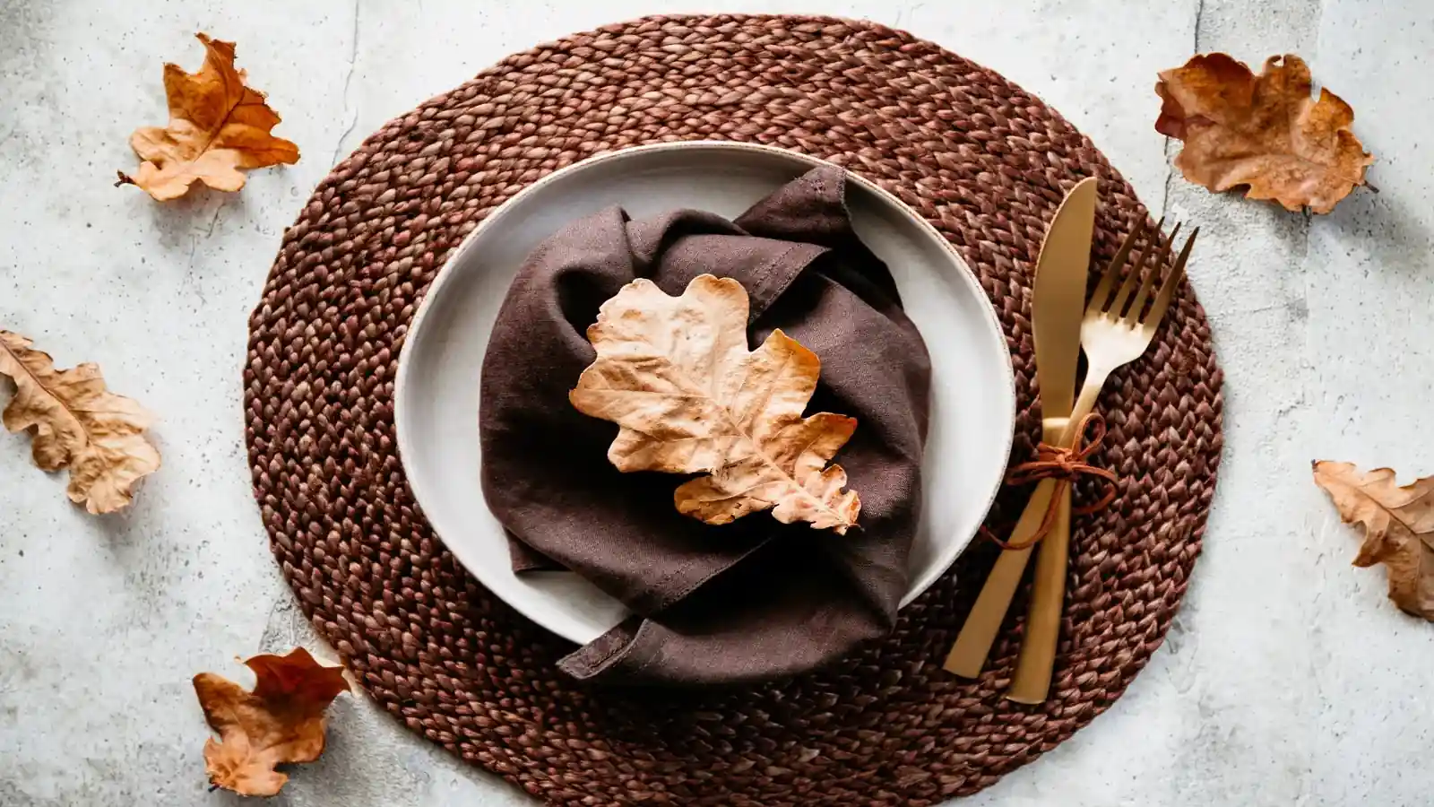 Elegant fall-themed table setting with a brown woven placemat, neutral plate, folded napkin, and dried leaves, creating a warm brown color party aesthetic