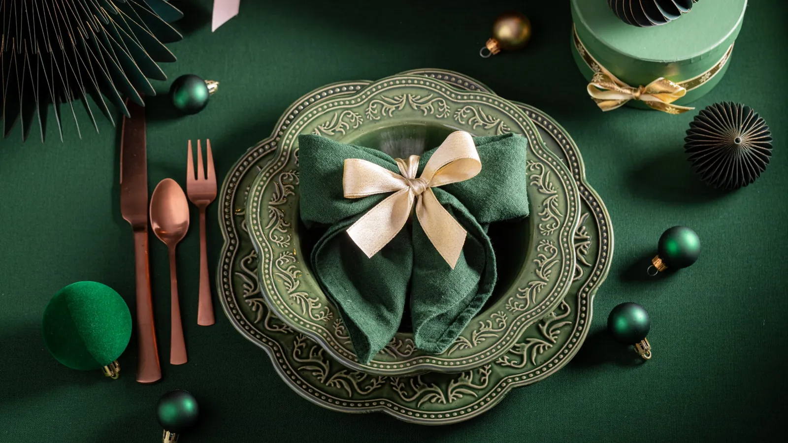 Elegant green-themed party table setting with decorative plates, folded napkin tied with ribbon, and holiday ornaments on a green background