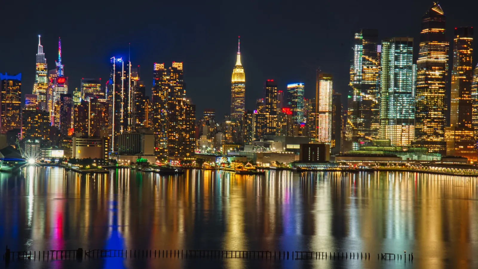 New York City skyline at night with illuminated skyscrapers and colorful reflections across the Hudson River