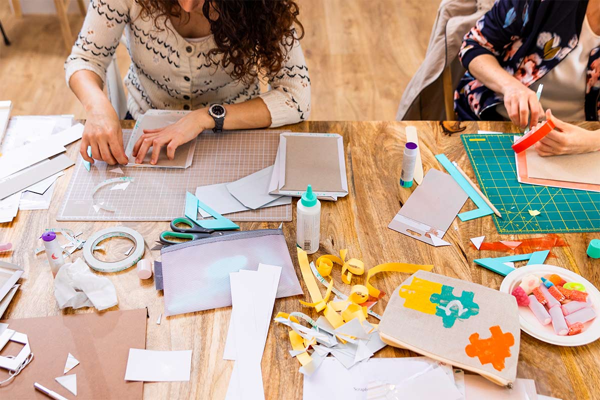 overhead view of two women sitting at a wooden table full of crafting supplies and making scrapbook pages