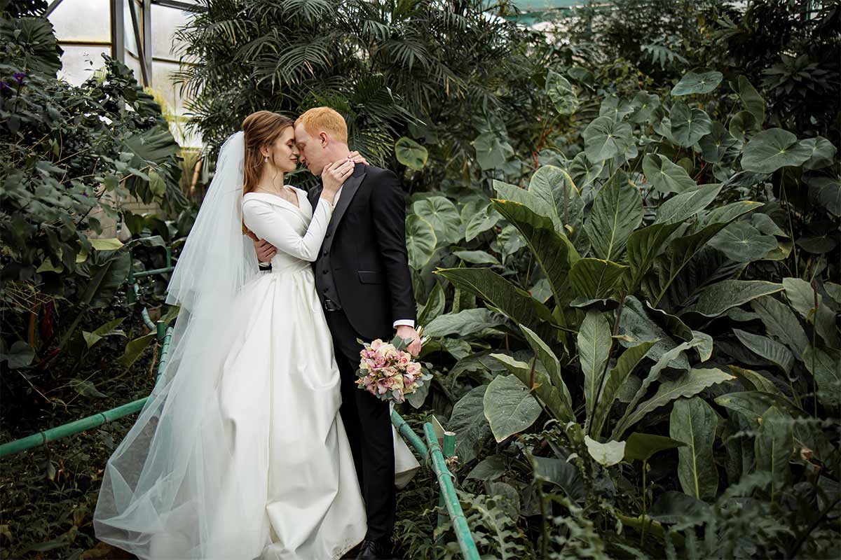 White 25-year-old bride wearing a white wedding dress and veil embracing her 25-year-old white groom in a botanical garden in a greenhouse. 