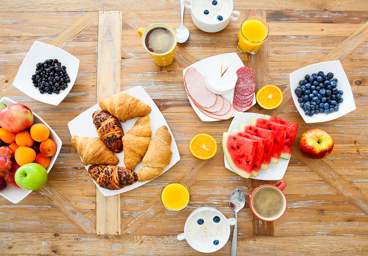 wooden table with a breakfast spread that includes white plates with blueberries, watermelon, croissants, meats, and other fruits also there are glasses of orange juice and cereal bowls