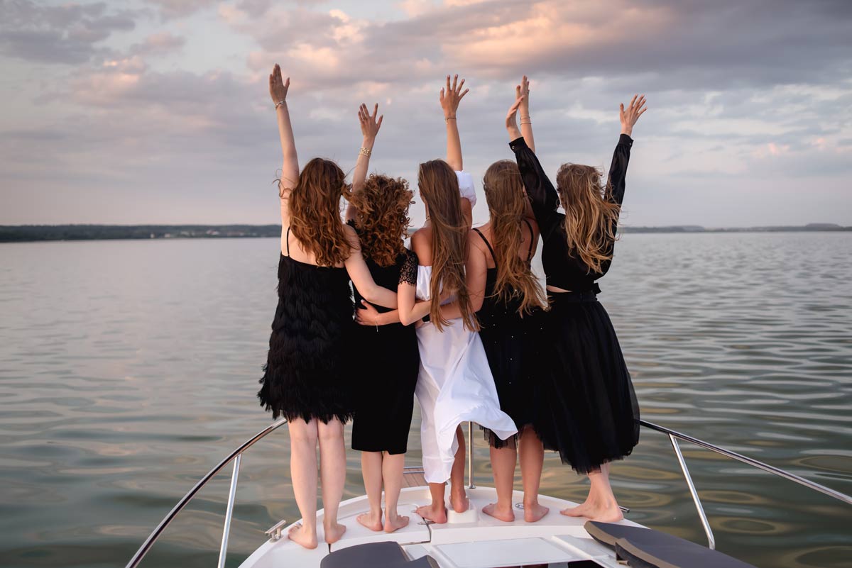 Group of women in elegant long dresses, with the bridal party in black dresses and the bride in a white dress, standing at the front of a boat in the middle of the water facing away from the camera with hands in the air.