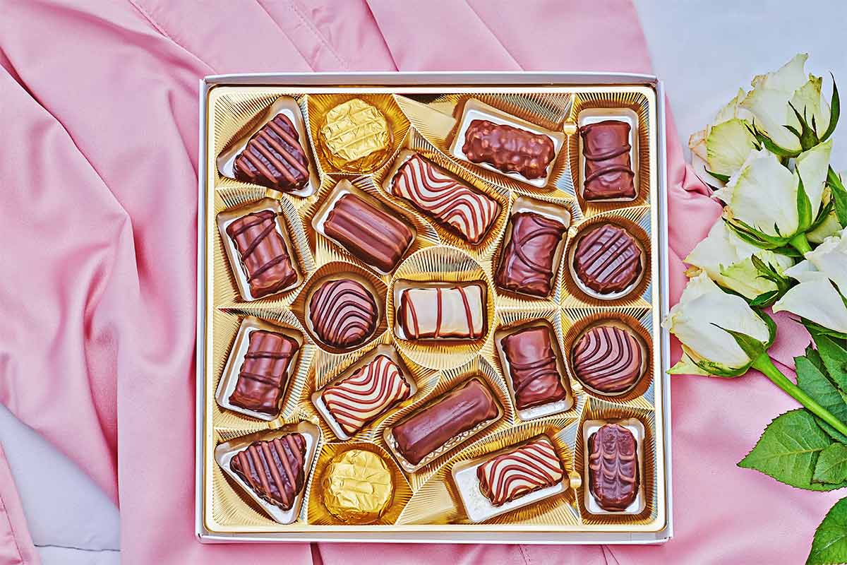 Assortment of chocolates that are placed on top of a pink fabric and next to white roses. 