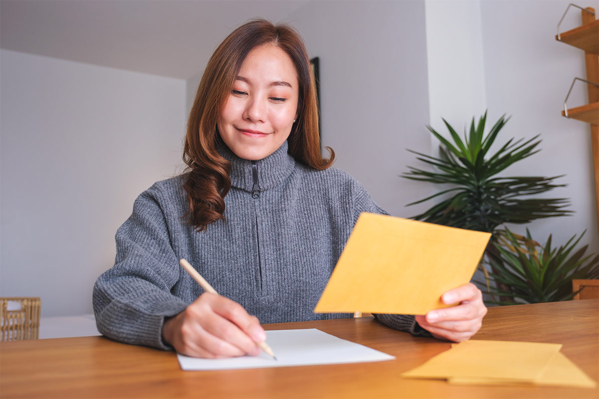 Asian woman in her 30s wearing a gray sweater sits at a wooden desk and smiles as she writes on a piece of white paper and holds a yellow envelope