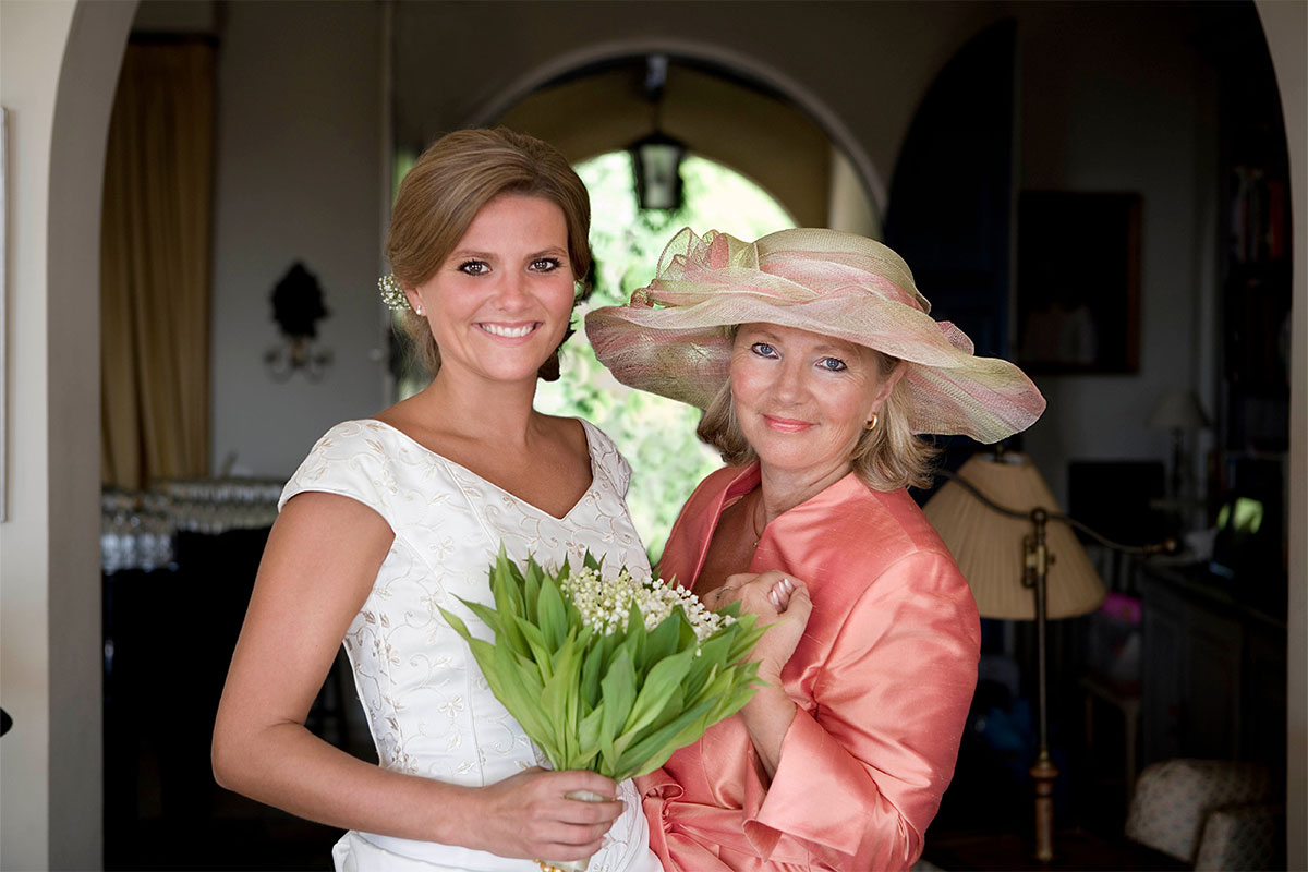 A white early 40s bride holding a bouquet and posing with her mother who wears a coral pink dress and large hat