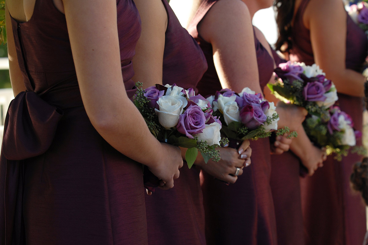 A row of multiracial bridesmaids in deep purple dresses holding white and purple bouquets