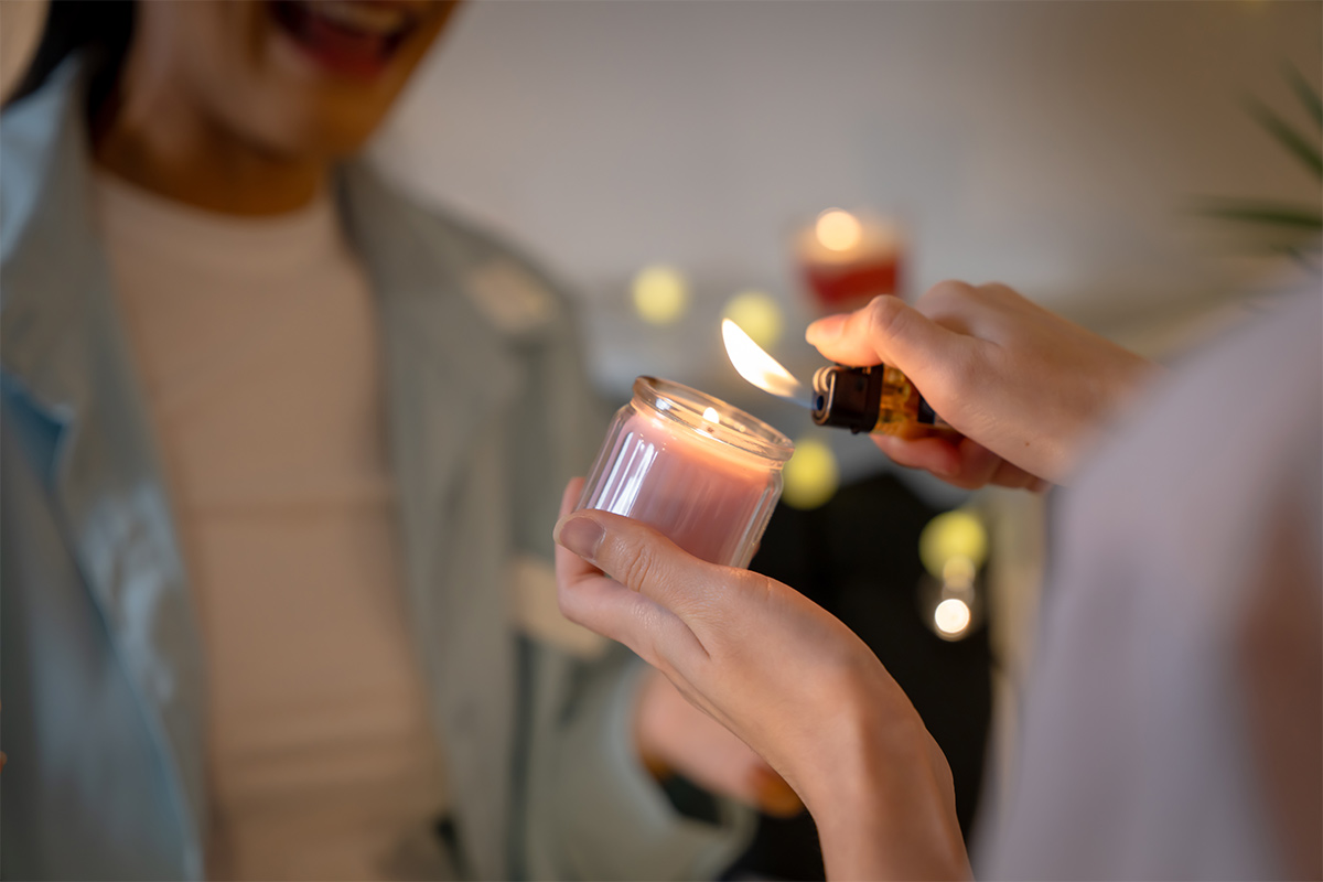 one asian woman lights a pink candle in a jar with a lighter while another asian woman wearing a blue shirt looks on excitedly 