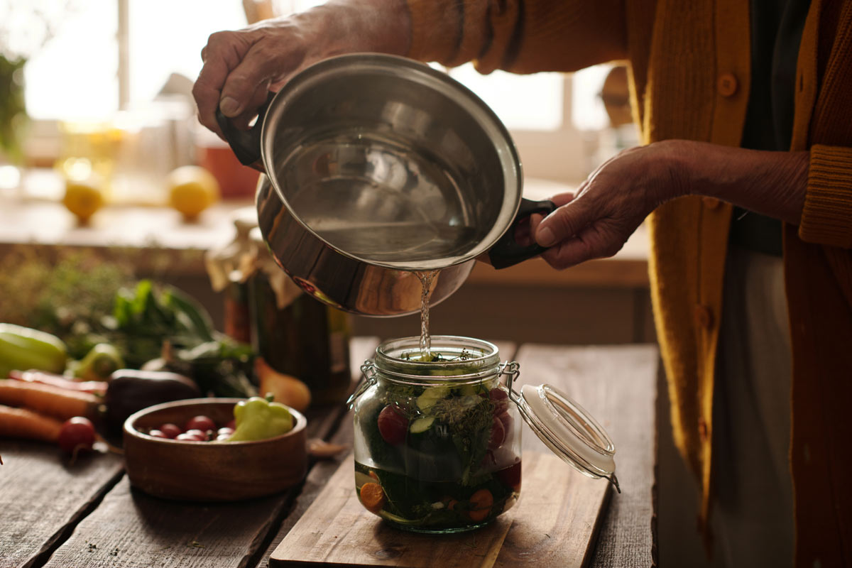 Close-up of a woman’s hands pouring hot water into a canning jar filled with vegetables, showing the process of home canning to preserve food.