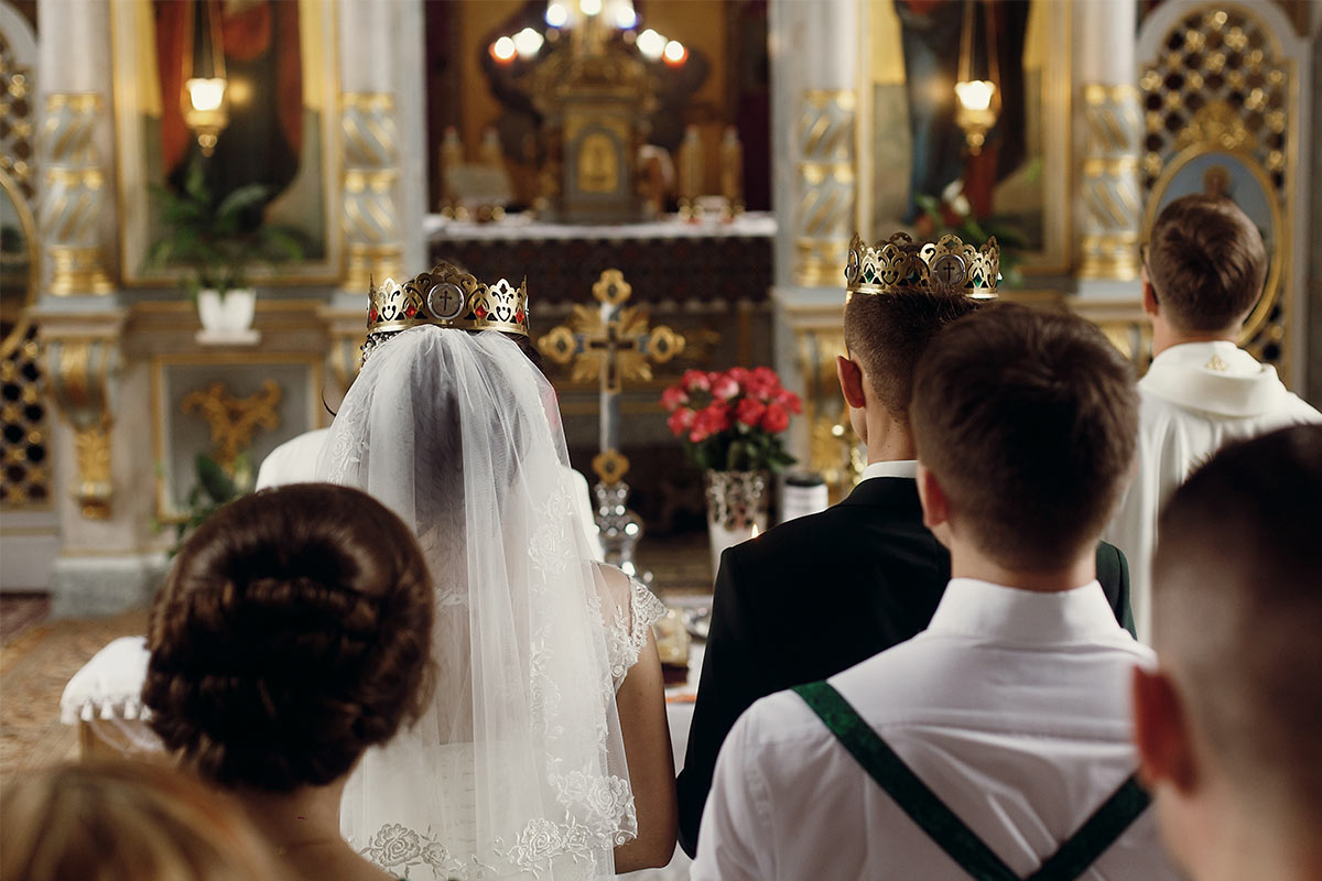 A bride and groom wearing religious crowns with their wedding party kneeling at the altar in a church