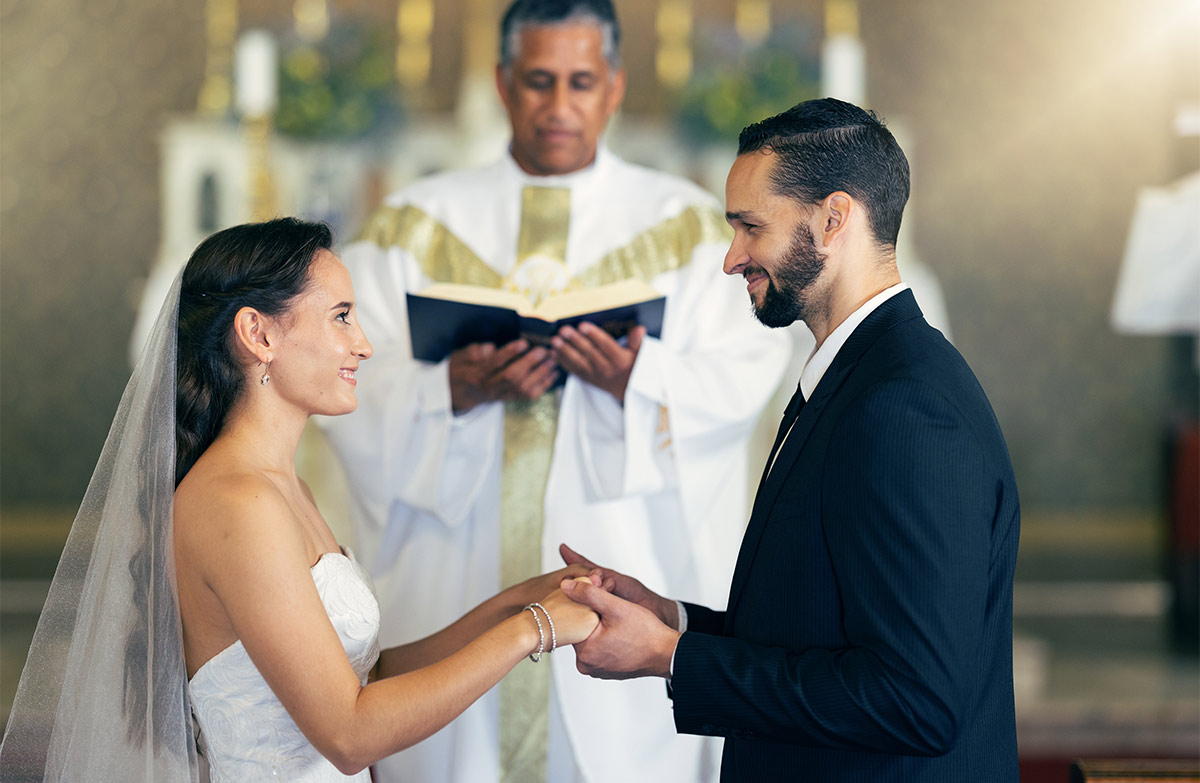 A 30-something white bride and groom holding hands at an altar with priest reading from bible behind them