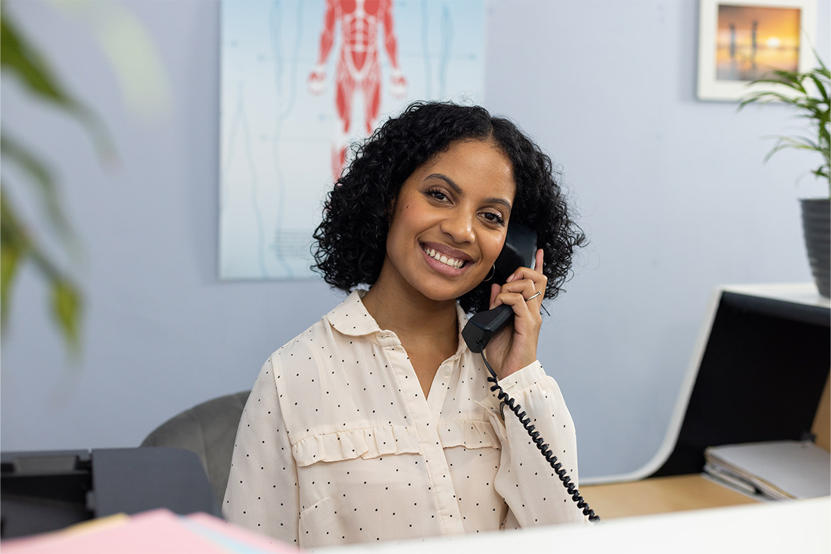 Black woman receptionist smiling while answering the phone while sitting at her desk at a doctor’s office.