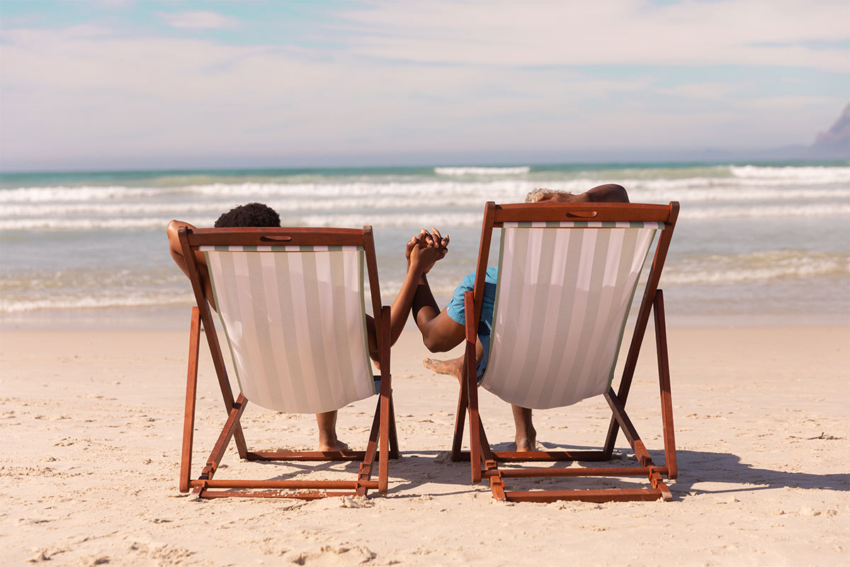 Black couple holding hands in beach chairs on the sand facing the ocean.