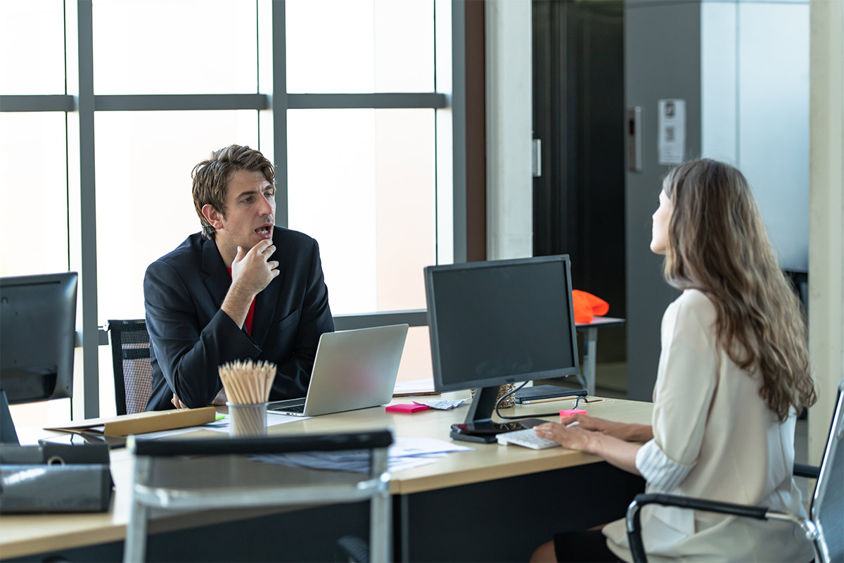 White man and white woman having a conversation with each other at a desk with computers in front of them in an office.