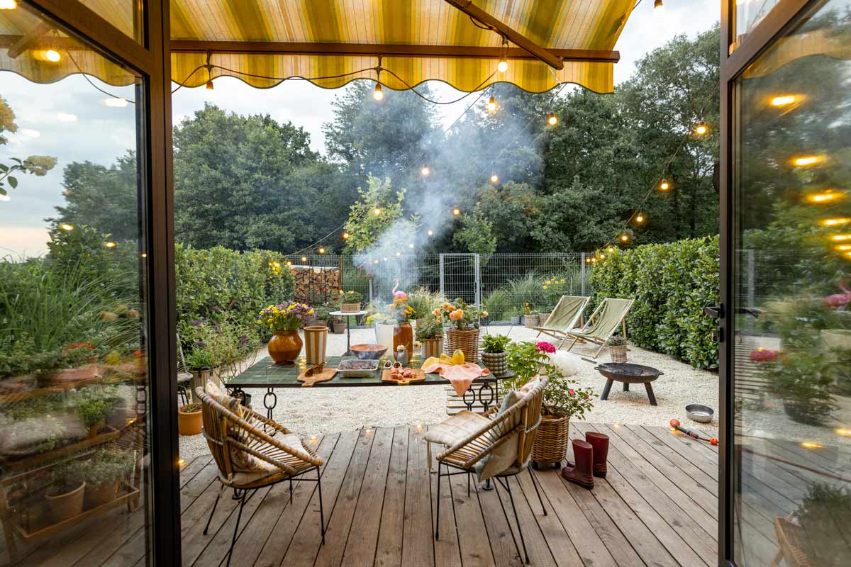 Backyard patio outside, with furniture, plants and a bonfire, with background lined with trees and shrubs. 