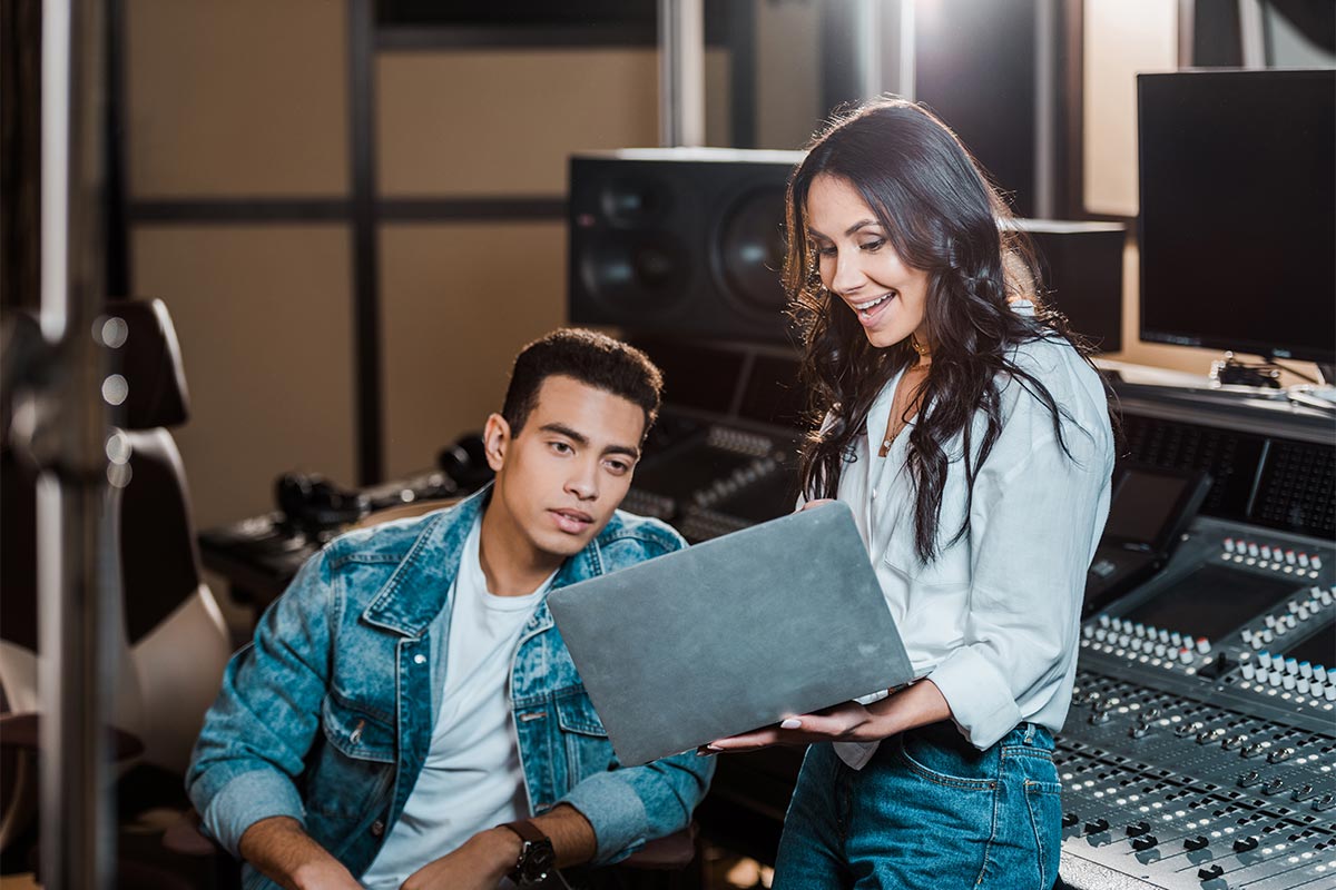 A smiling white woman in her 30s engaging with a sitting young black man in his 20s, pointing out information on a laptop in a studio setting