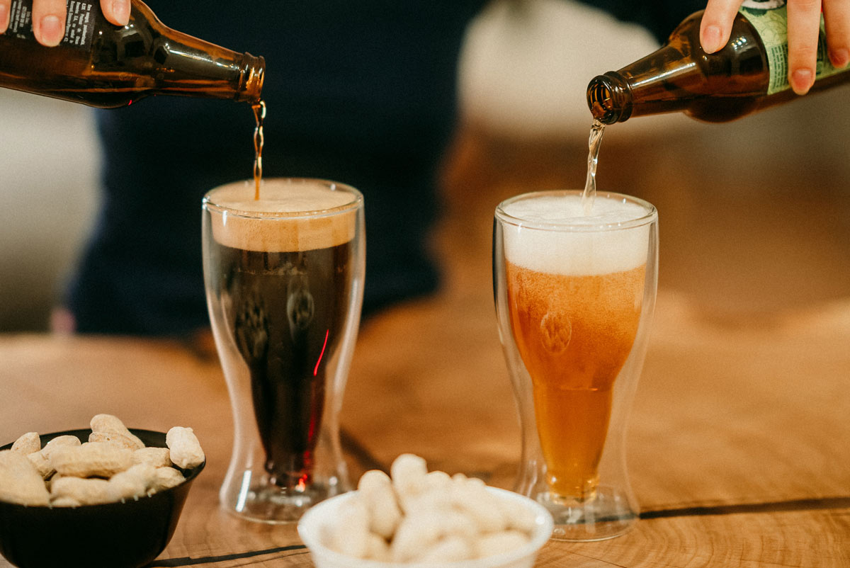 White person's hands pouring two beer bottles into glasses on a wooden table. One beer is darker than the other. 