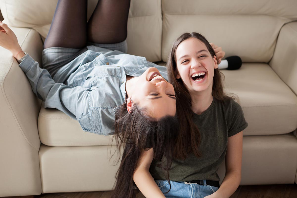 White teen sisters laughing together - one is upside down on the couch and one is seated on the floor in front of it - with a microphone in the background