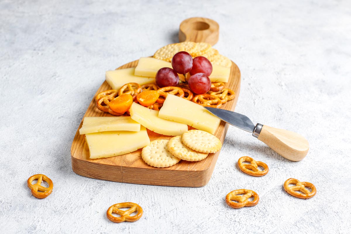 Cheese, crackers, pretzels, and grapes on a wooden cutting board, with a little knife next to it, on  a white table