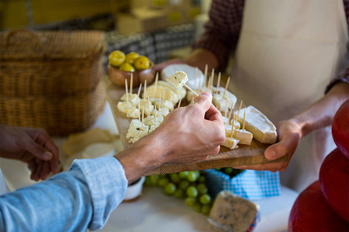 White hand reaches grabs a toothpick with cheese off of a wooden board of assorted cheeses 