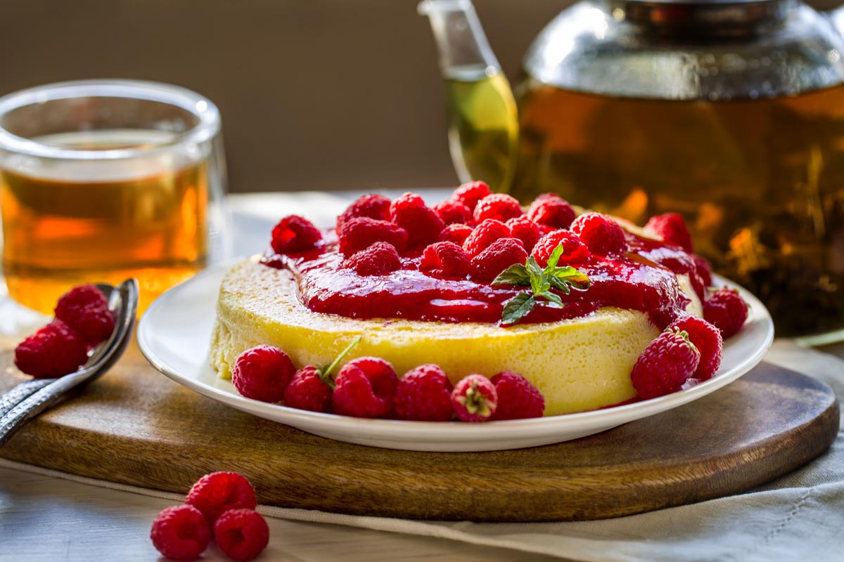 Cheesecake slice topped with fresh raspberries and raspberry jam on a wooden kitchen board with tea behind the cheesecake in a clear glass cup and glass teapot, offering a balanced dessert with a fruity twist.