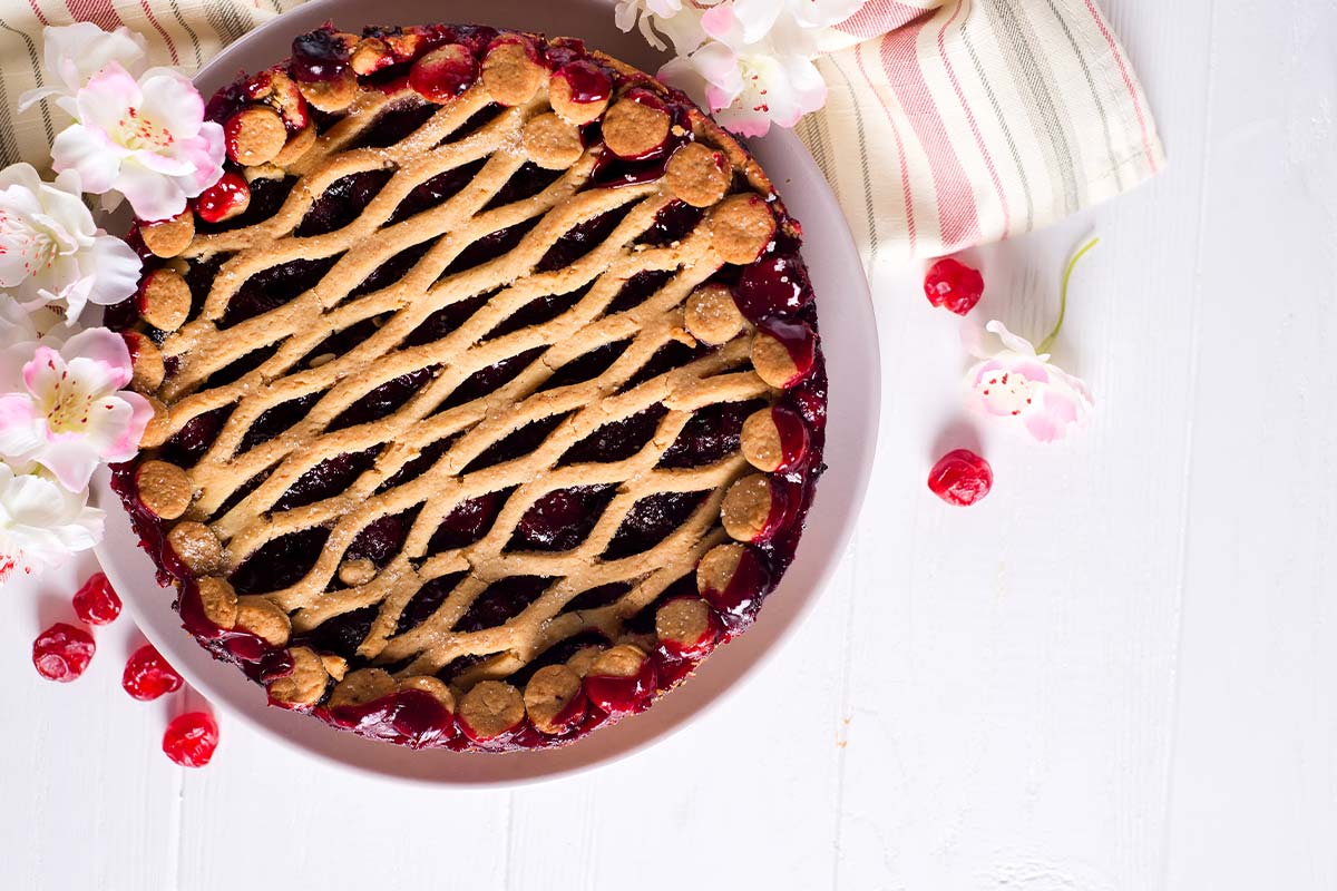 Birdseye view of a cherry pie in a white pie dish surrounded by loose cherries and blossom flowers.