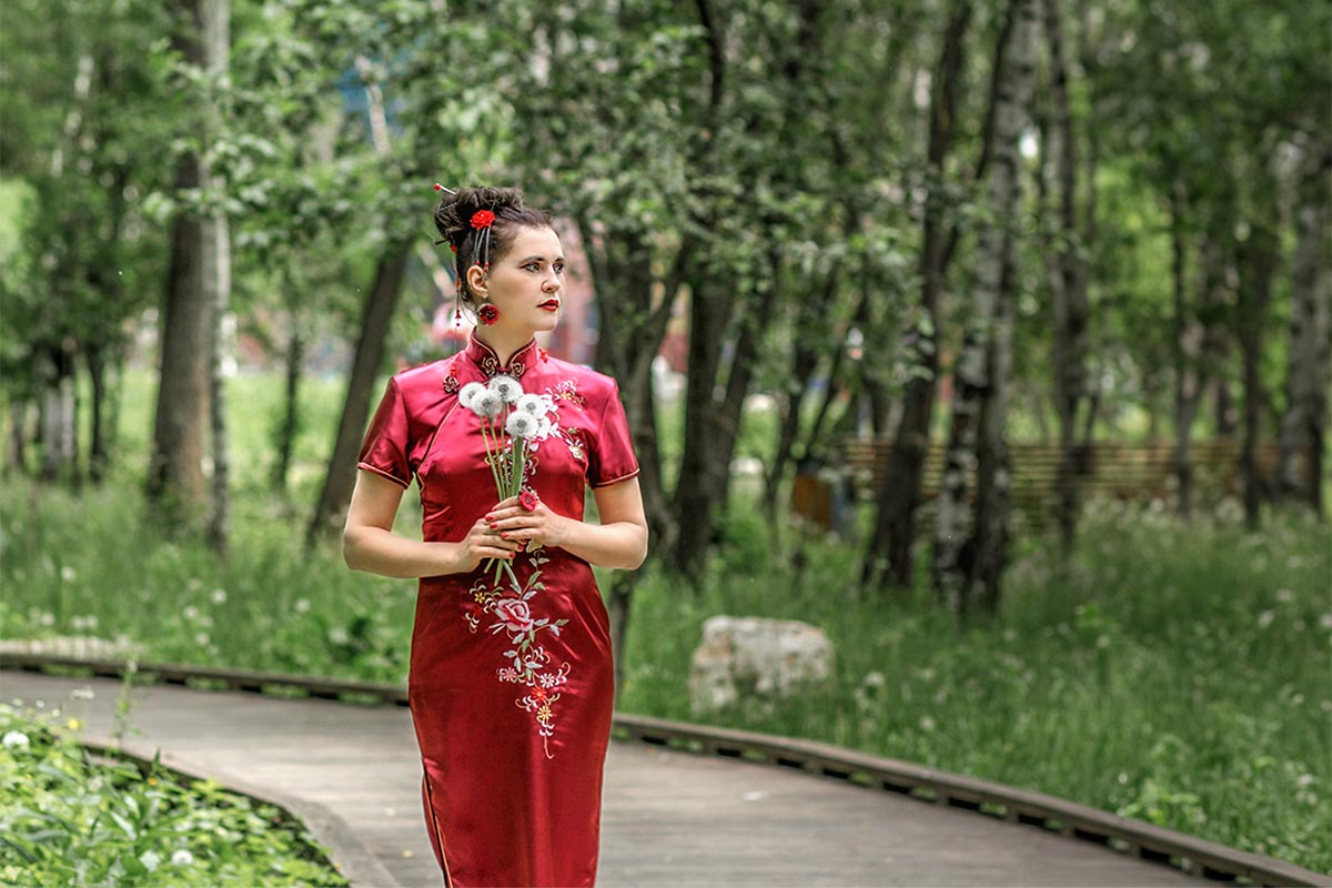 30 something Asian woman in traditional red Asian wedding dress holding bouquet of dandelions and looking in distance while walking on outdoor path surrounded by trees and grass