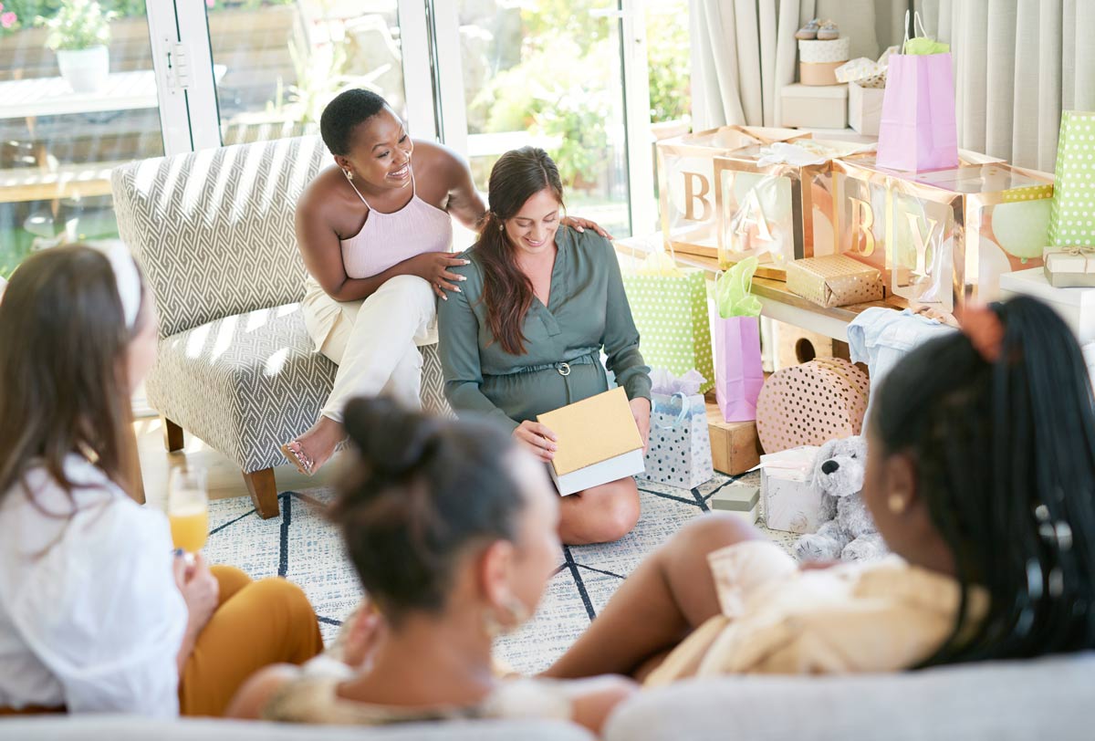 30 something pregnant white women sitting down on the living room floor opening up baby shower gifts, with her partner behind her, while friends and family are watching with their backs turned away from the camera. 